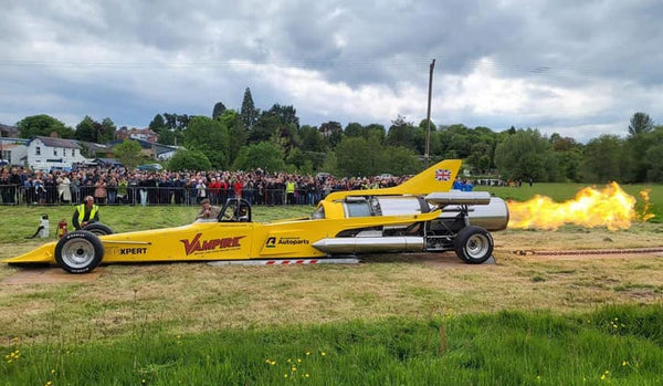 The Vampire Jet vehicle featuring the Arnold Clark Autoparts logo shoots a large orange flame from its rear. In the background a crowd behind a barrier watch on at the Bromyard Speed Festival.