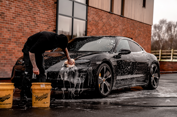 Man with a microfibre cloth in hand washing his black Porsche outside a brick building. He is dressed in black with two yellow Meguiar's 5 gallon buckets at his feet.