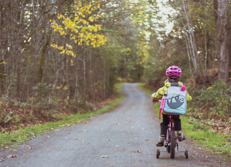 young girl wearing a colourful helmet, coat and backpack, cycles alone on an empty path. She is surrounded by autumnal forestry and falling leaves. 